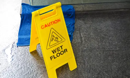 A yellow warning sign next to a puddle and a wet rug on a stone floor.