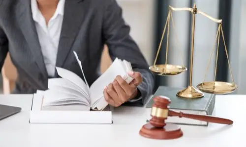 A close-up of a lawyer reading a law book at a desk, with a gavel and scales of justice placed nearby.